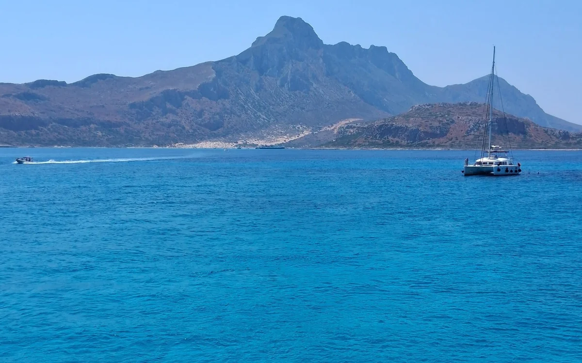 Bare peaks above deep blue sea, a sailing catamaran on the water and a speedboat trailing a white wake