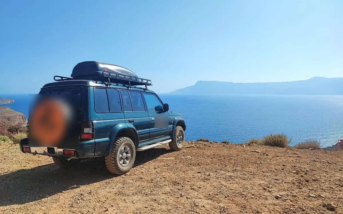 A dark green 4x4 parked on a dirt clifftop above open blue sea, with a long headland across the water