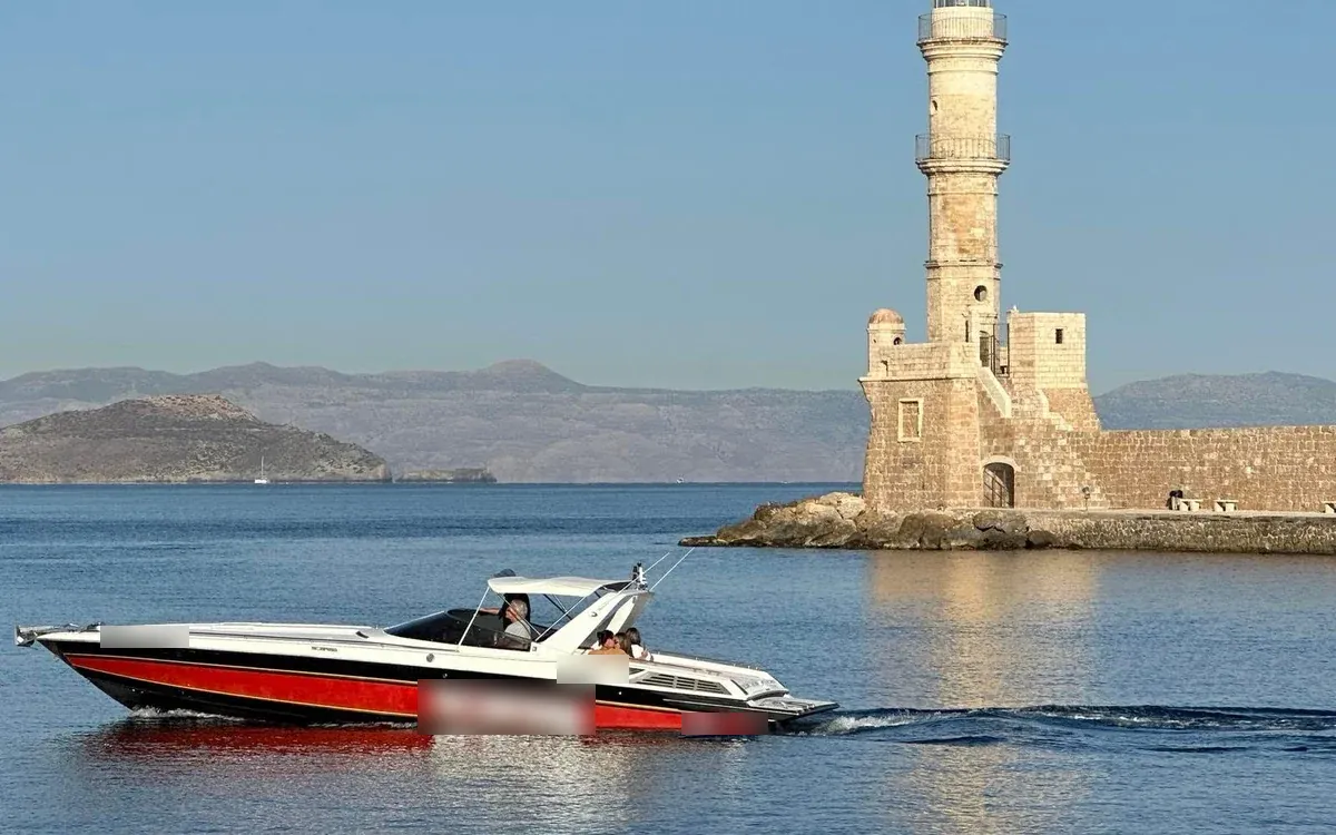 A red and white powerboat crossing calm water in Chania harbour, an old stone lighthouse behind it
