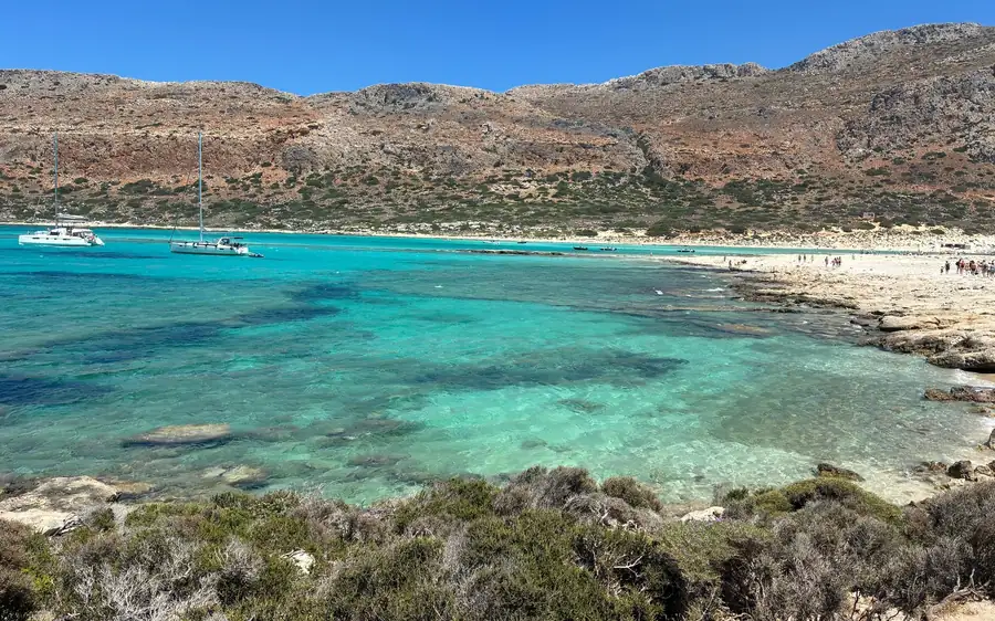 Turquoise shallows and a sandy shore seen from a scrubby hillside