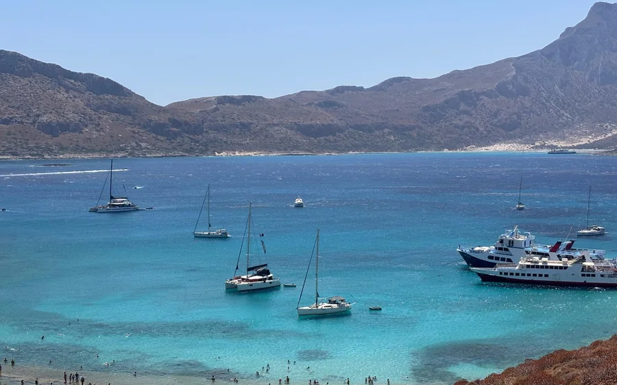 Sailing boats and a passenger boat moored in a turquoise bay below mountains