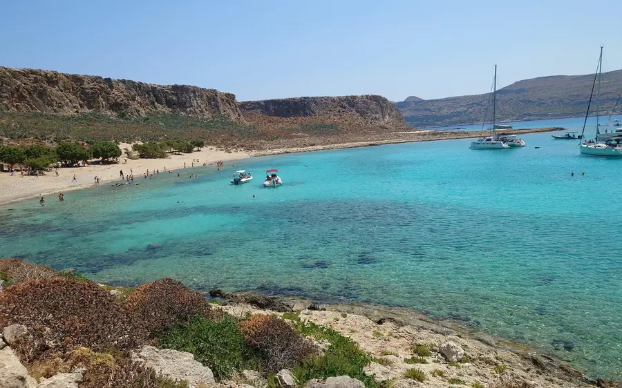 A turquoise bay below a headland, with sailing boats moored off a sandy shore