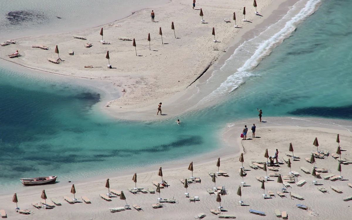 Rows of parasols and sunbeds on a pale sand bar between shallow turquoise channels, seen from above